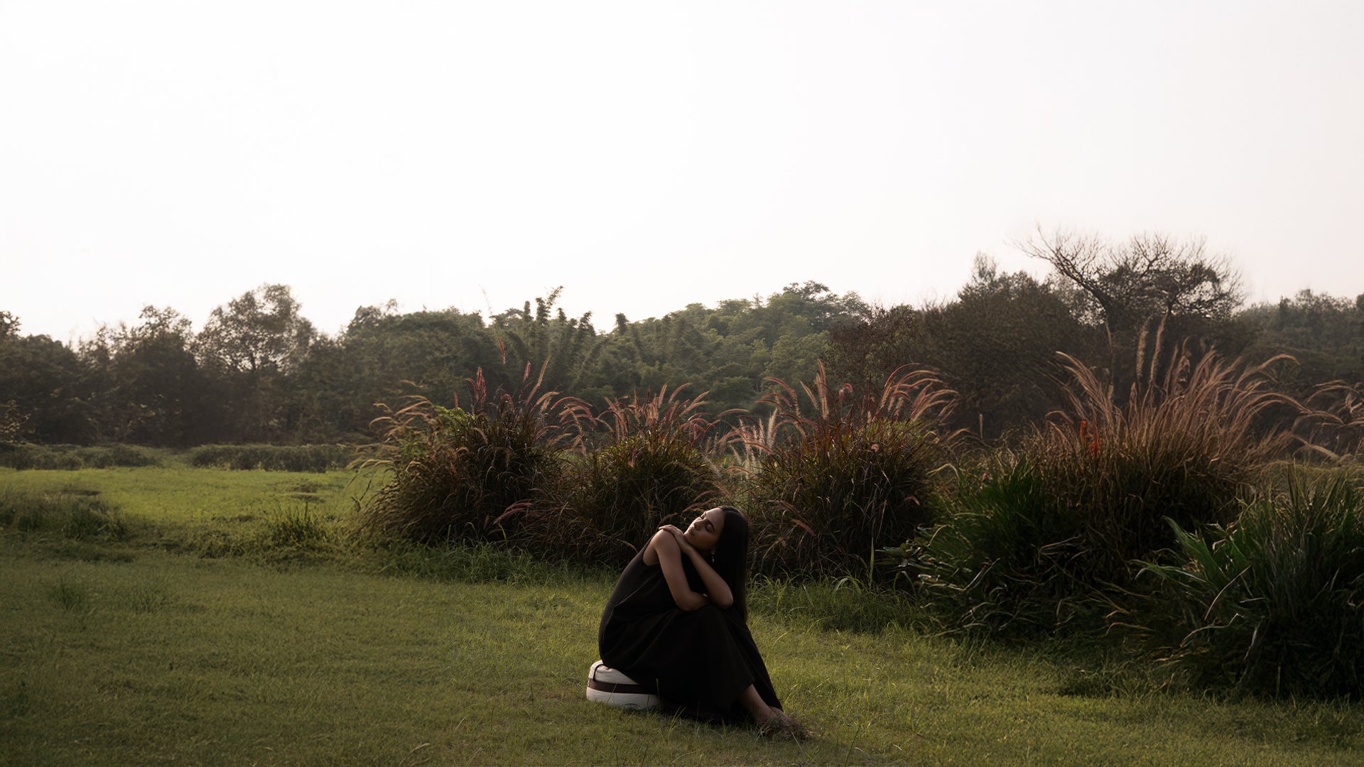 Person sitting on grass with arms crossed, surrounded by tall grass and trees